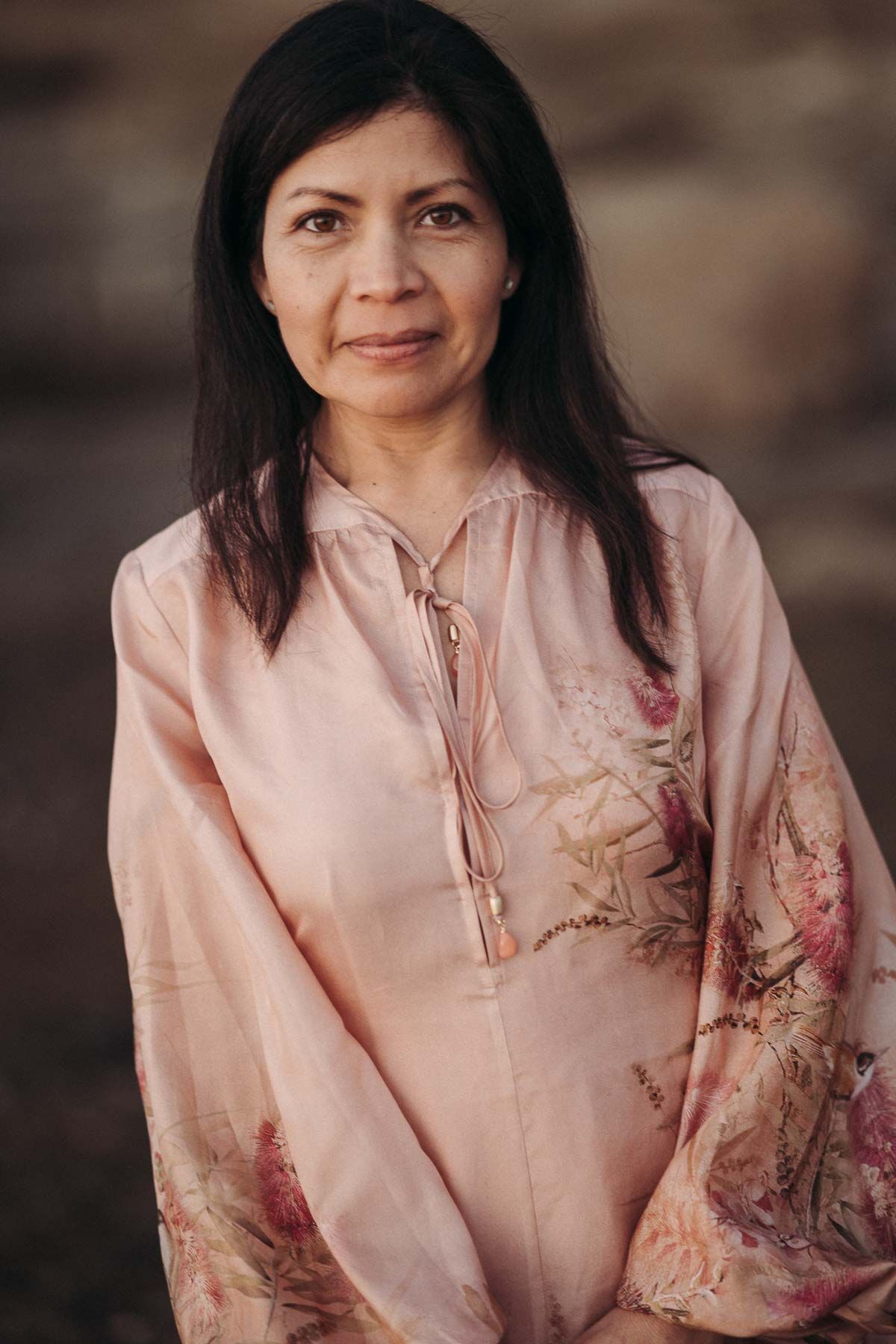 A person wearing a pink embroidered blouse with floral designs poses against a blurred background.