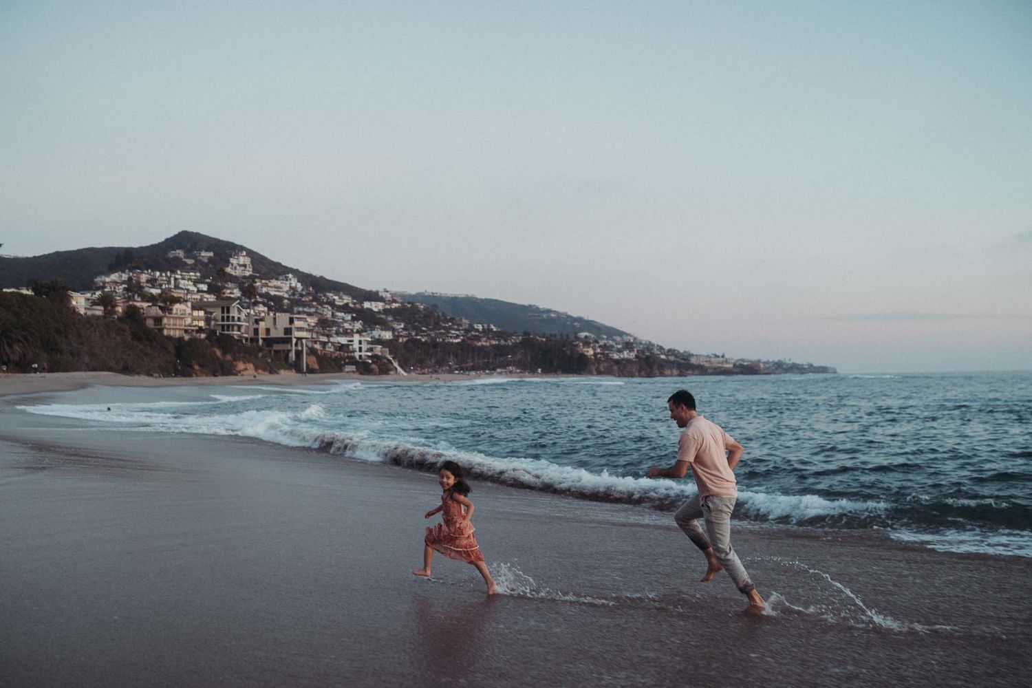 Two people playing and running along the shoreline at sunset with coastal buildings in the background.