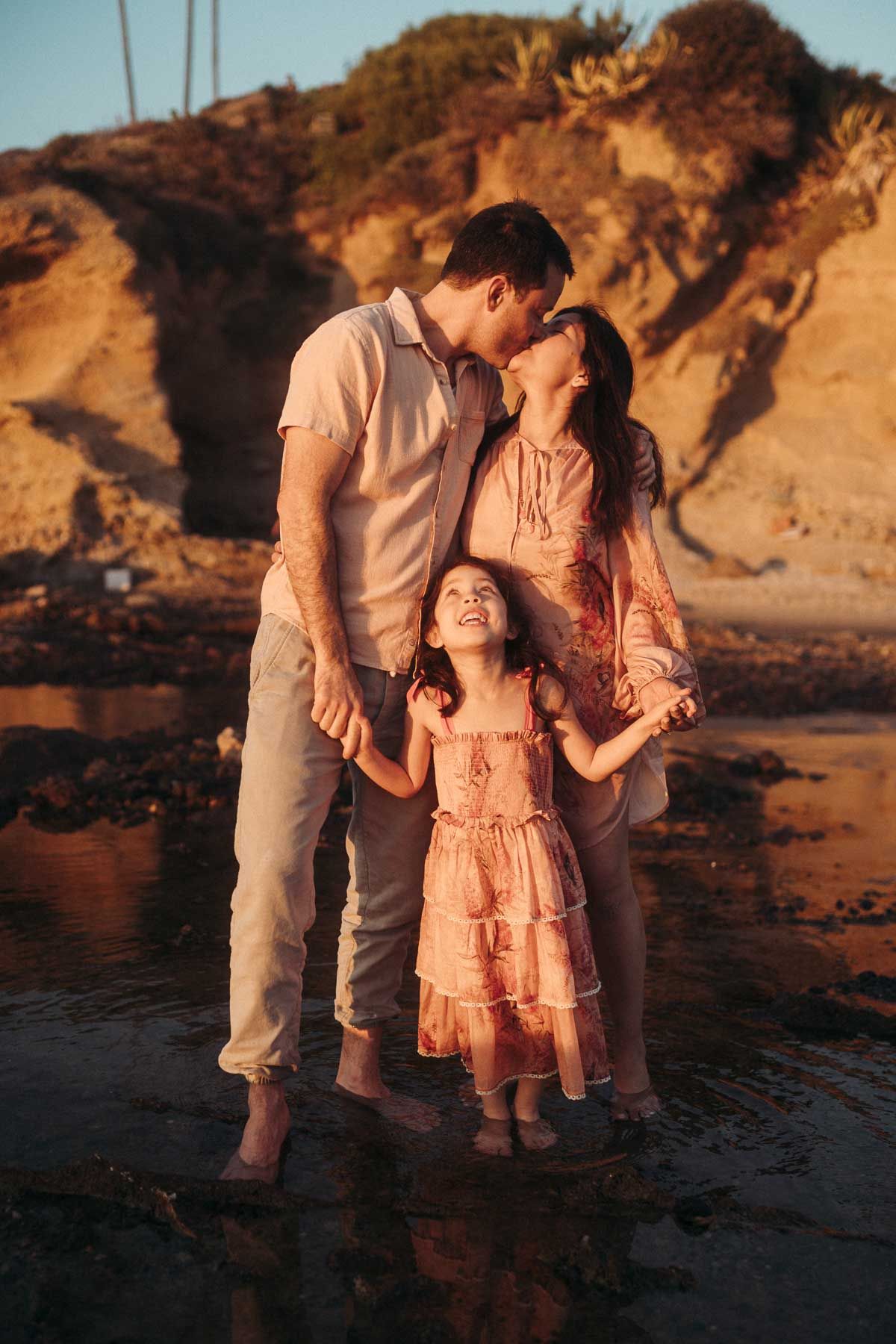 A family shares an intimate moment together at sunset on a beach with rocky cliffs in the background.