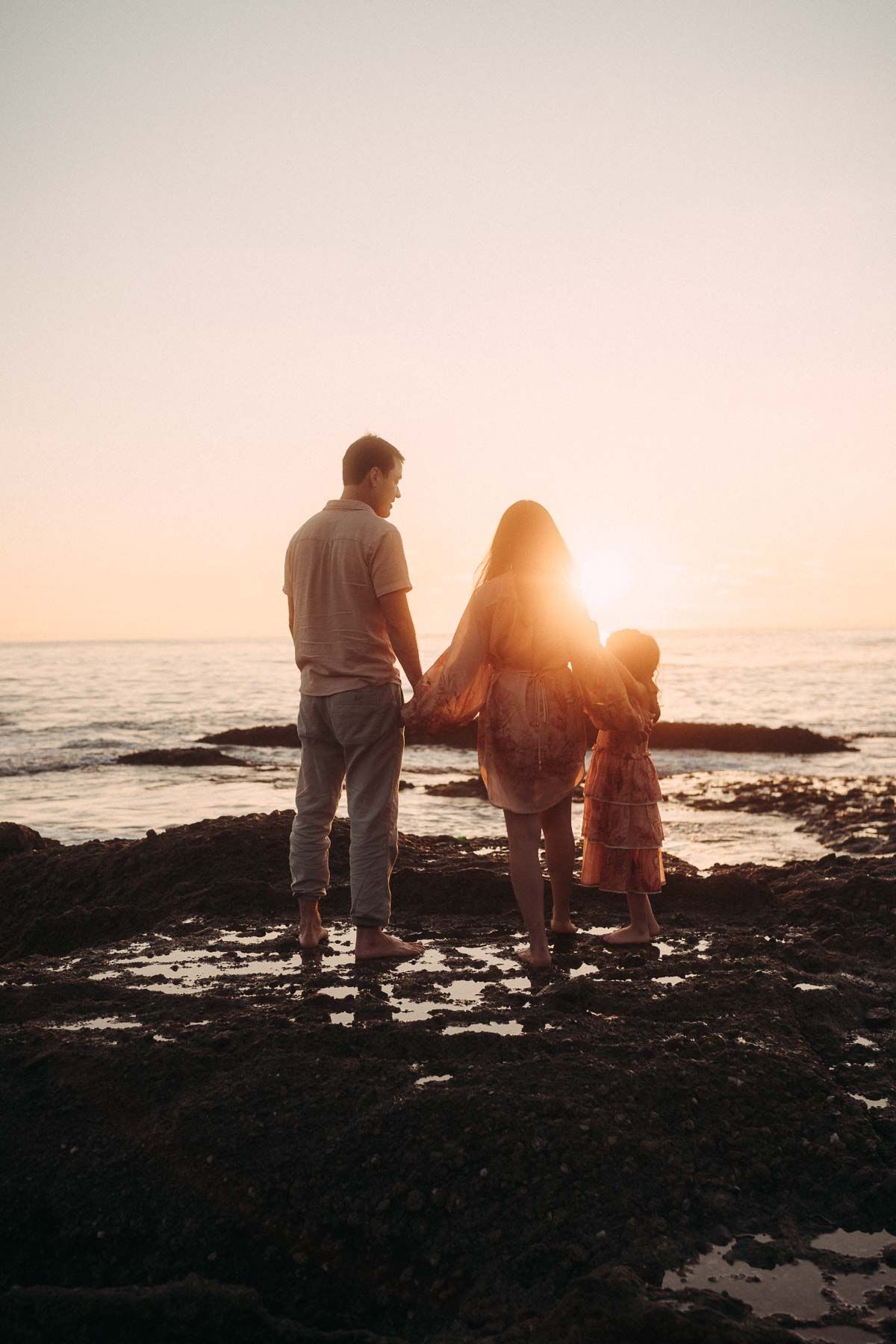 Three silhouettes stand on rocky coastline watching golden sunset over ocean horizon.