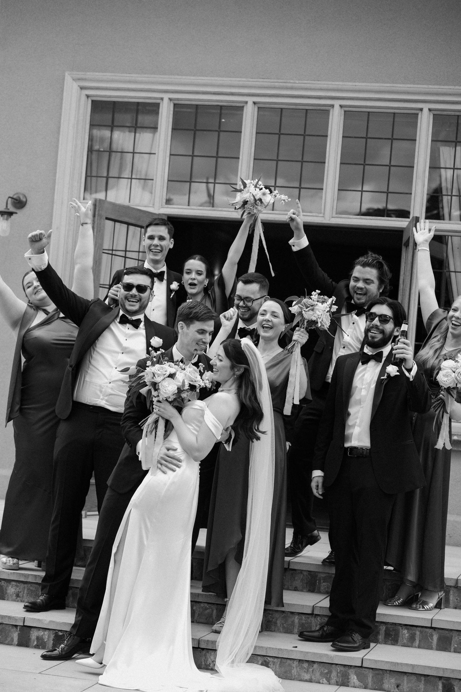 A joyful wedding party celebrates on steps outside, with the bride in a sleek white gown raising her bouquet in celebration.