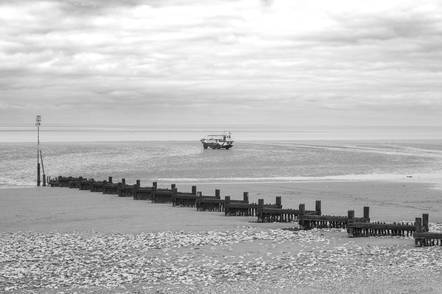 Black and white seascape showing wooden groynes extending into calm waters with a distant boat on the horizon.