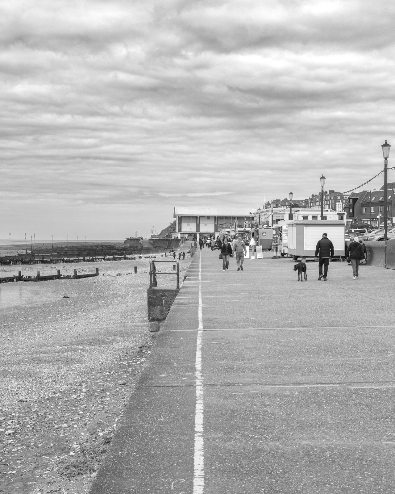 Black and white coastal promenade with people walking along a seaside path near beach and buildings.