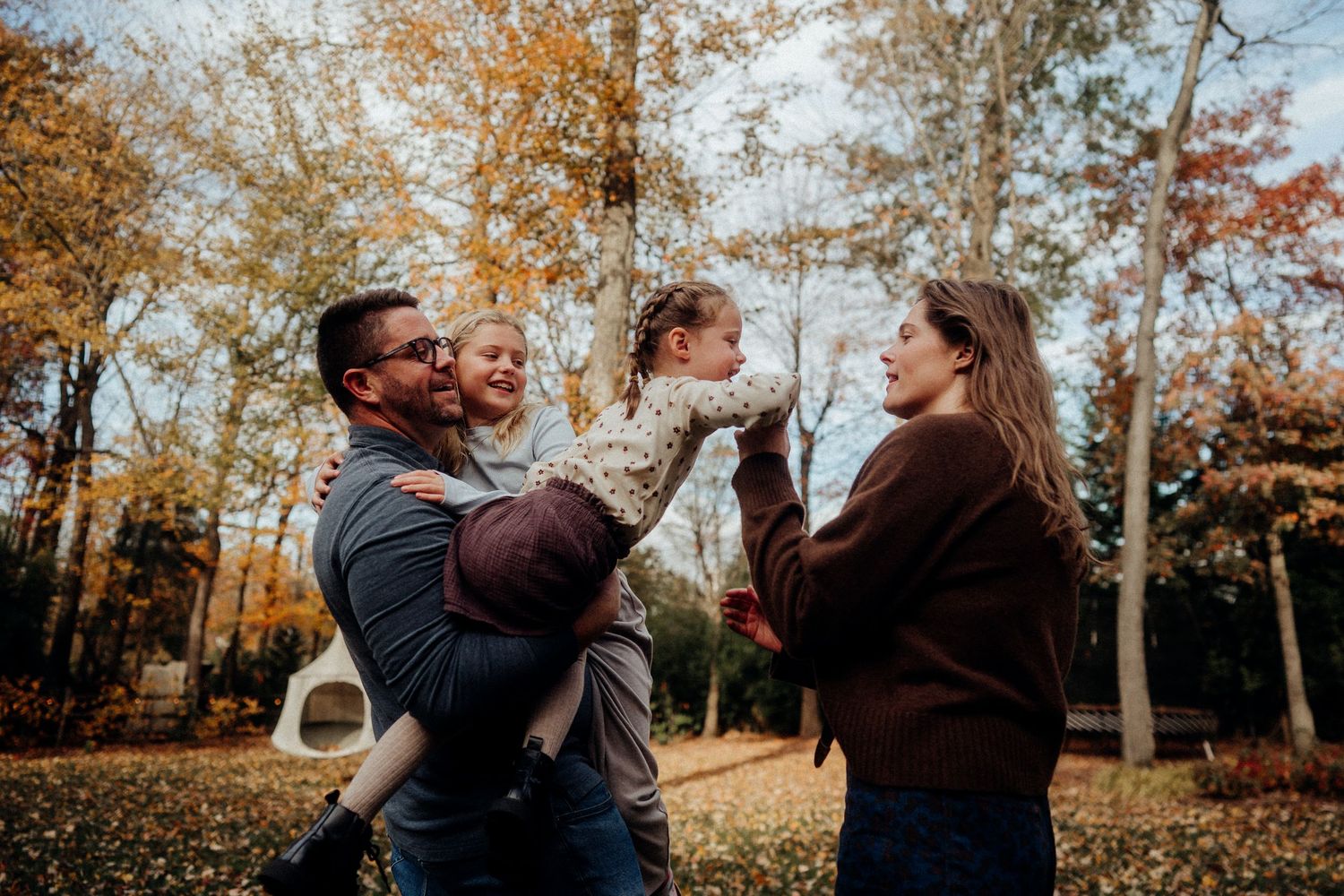 A couple plays with their small dog in a scenic autumn forest setting with golden leaves falling around them.