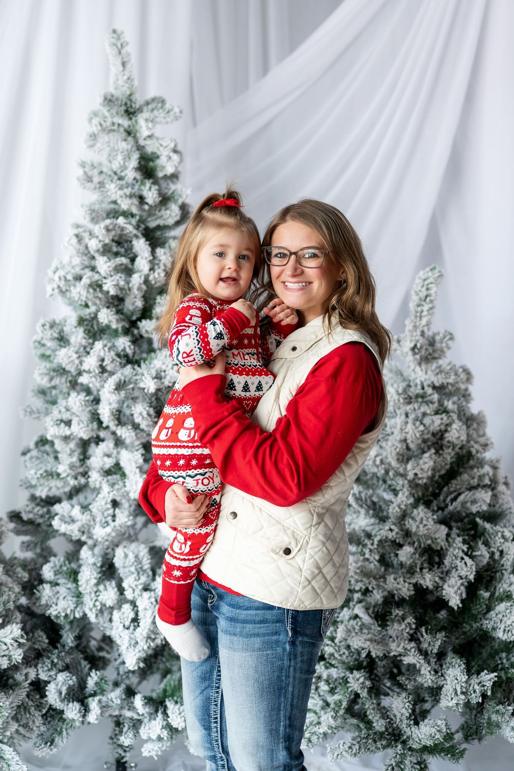 Smiling mother holding her young daughter in festive red Christmas pajamas in front of snow-flocked trees.