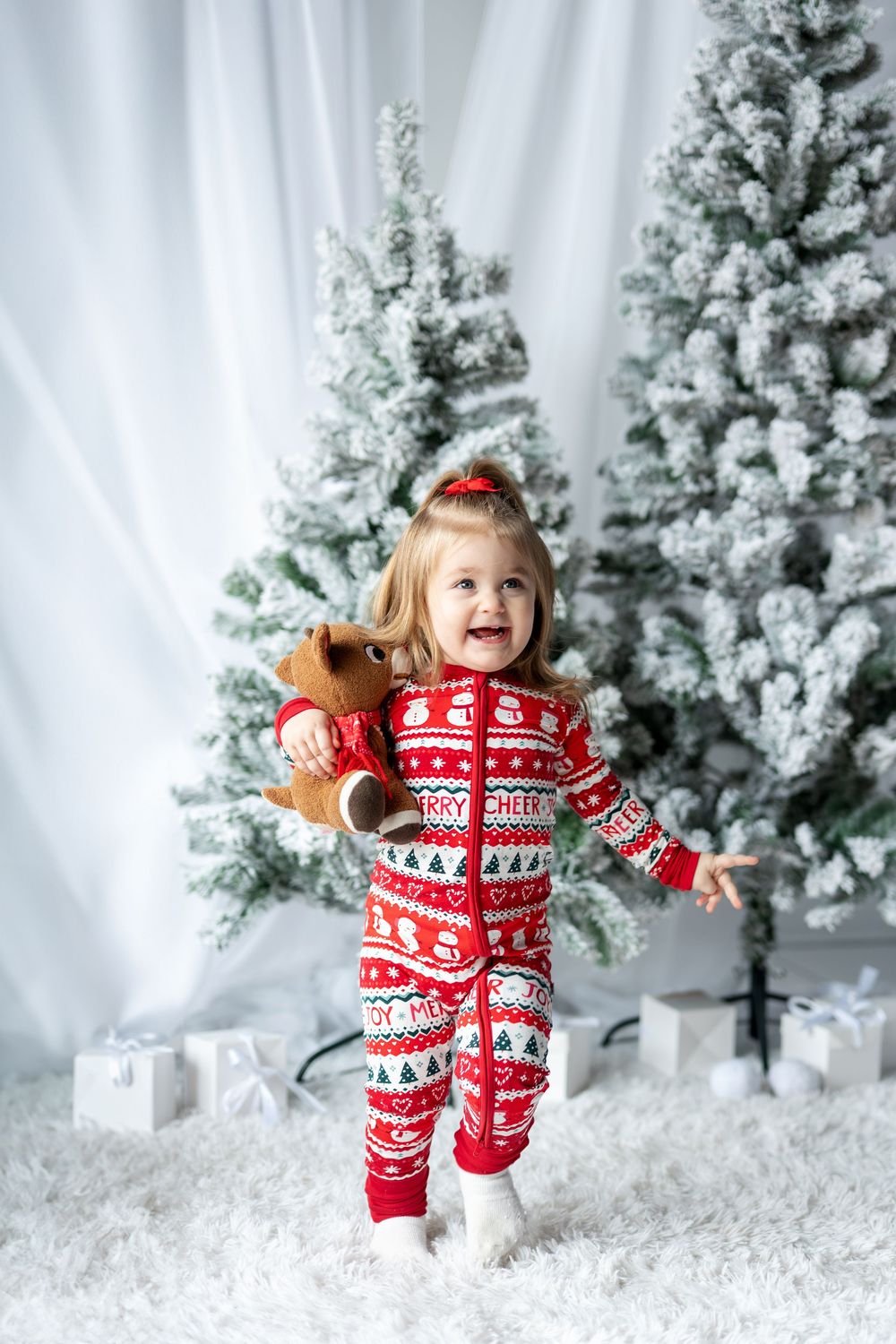 Joyful little girl in red holiday pajamas standing by snow-flocked Christmas trees.