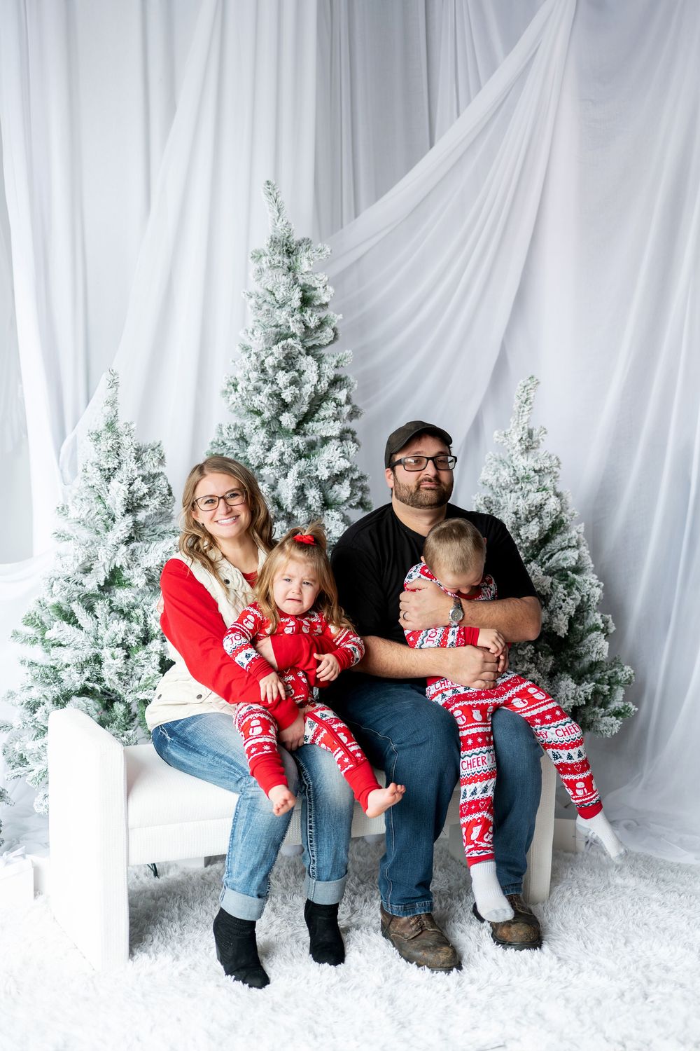 Family of four in Christmas pajamas sitting together by flocked holiday trees.
