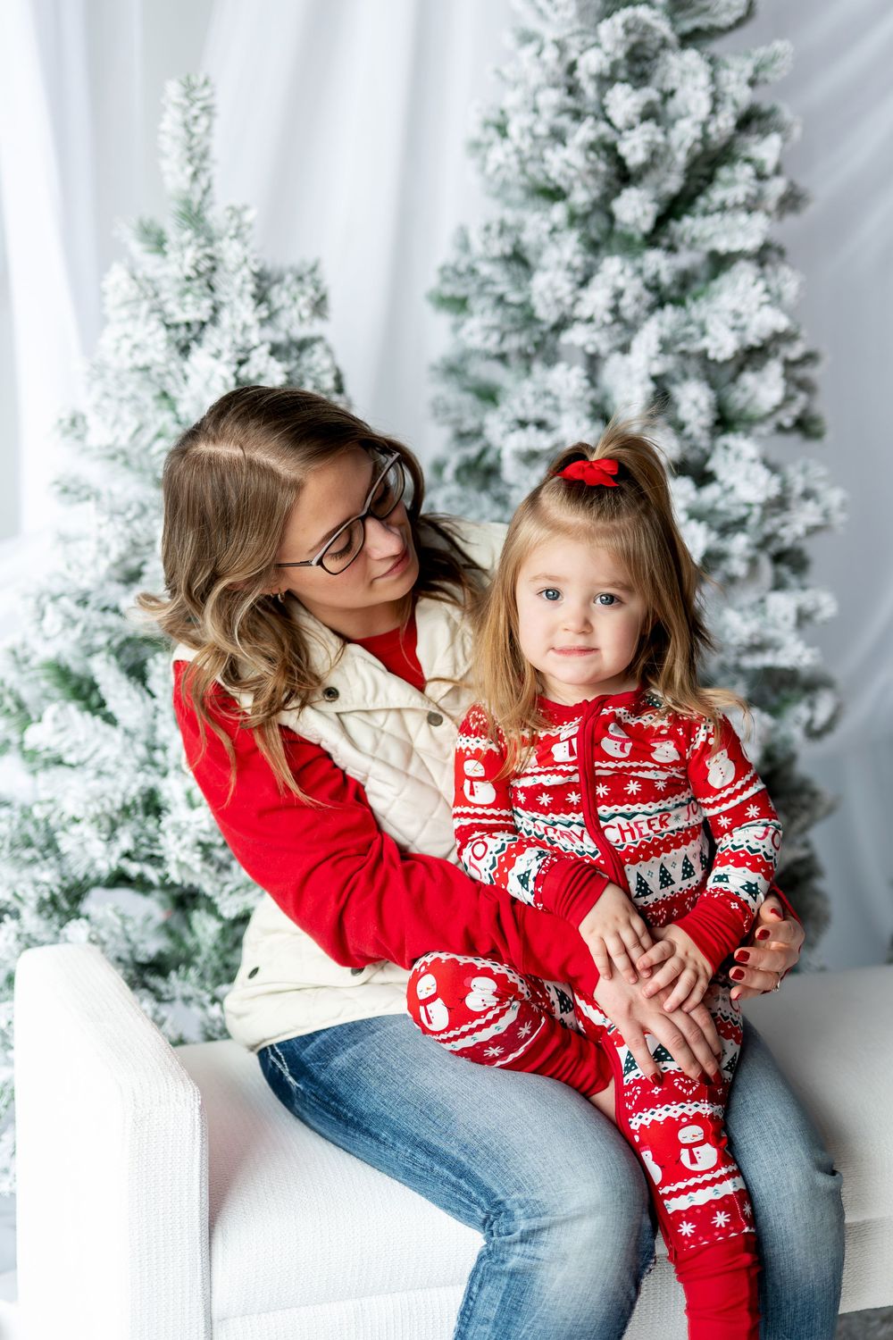Mother hugging daughter in red Christmas onesie sitting by snowy holiday trees.