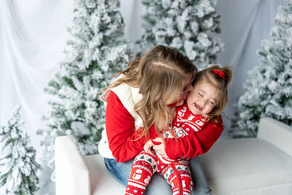 Mom cuddling laughing little girl in red Christmas pajamas on white bench.