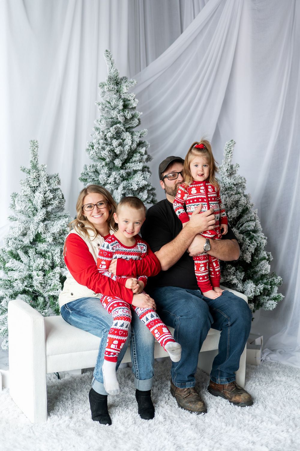 Happy family of four in matching Christmas pajamas posing by snow-flocked trees.
