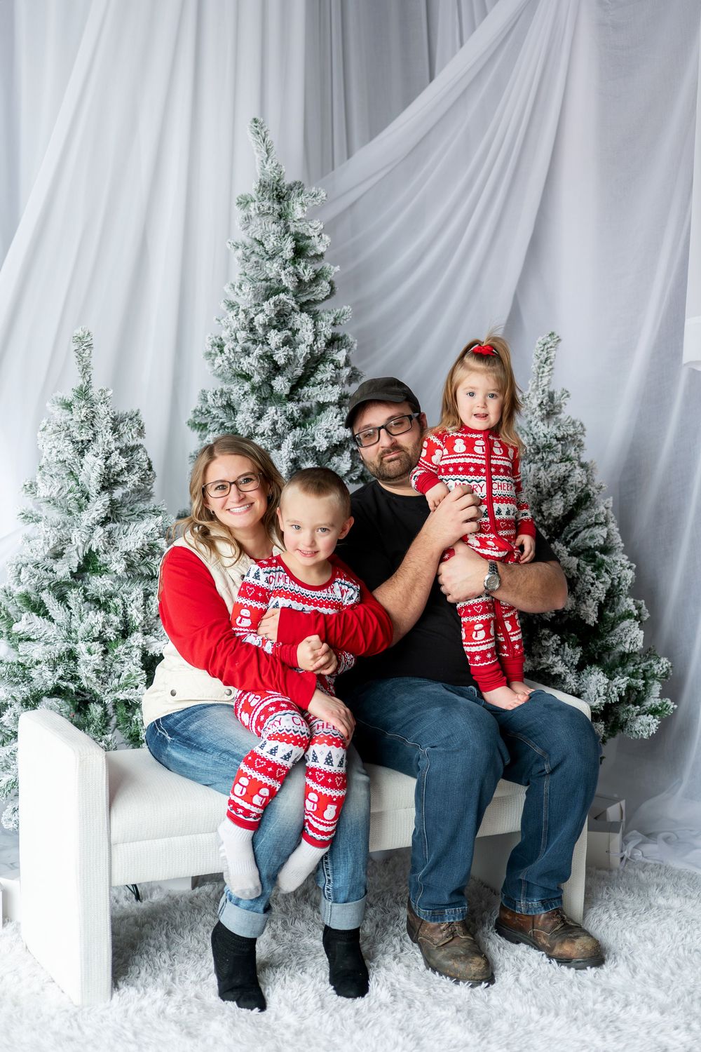 A cheerful family of four poses in matching red holiday pajamas beside snow-flocked Christmas trees.