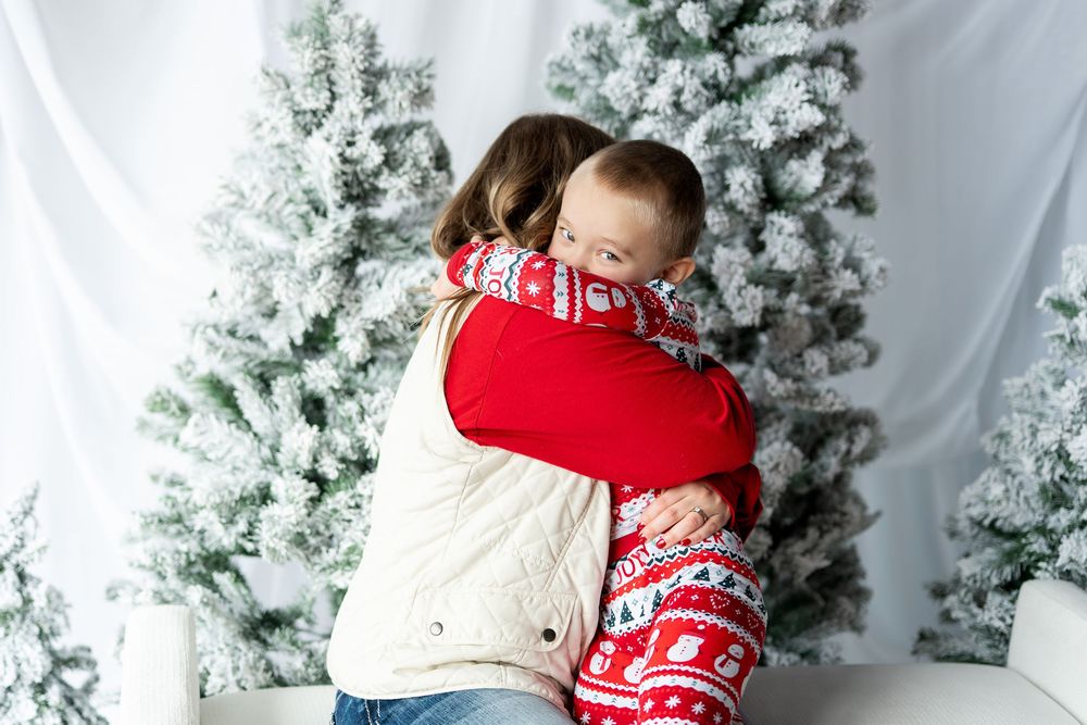 A mother tenderly embraces her young son in matching red Christmas pajamas near snowy holiday trees.