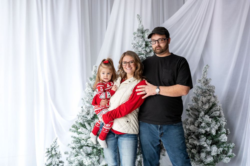 Family of three posing for a Christmas portrait with snow-flocked trees, mother holding young daughter.