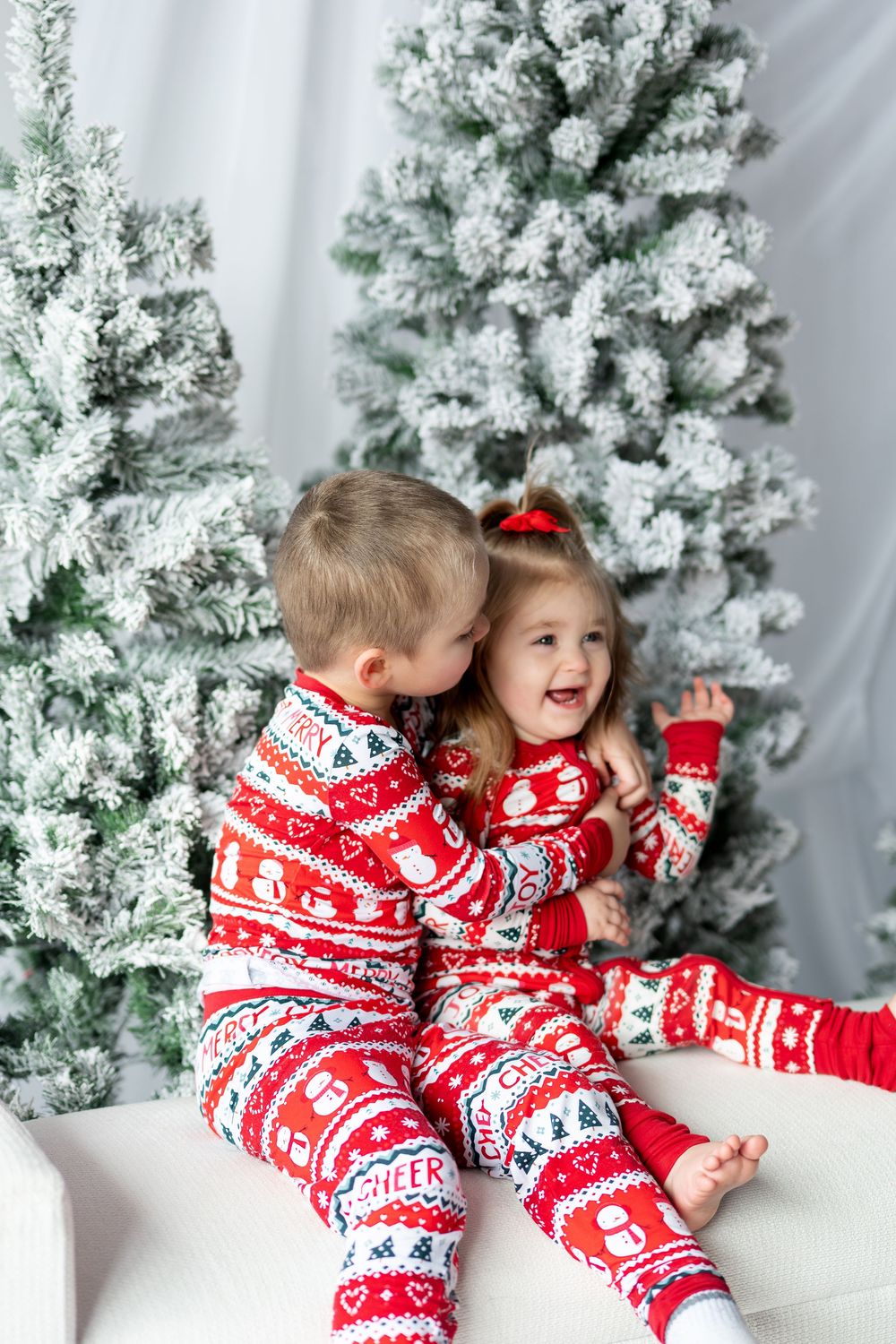 A young boy whispers to his laughing little sister, both wearing matching red Christmas pajamas.