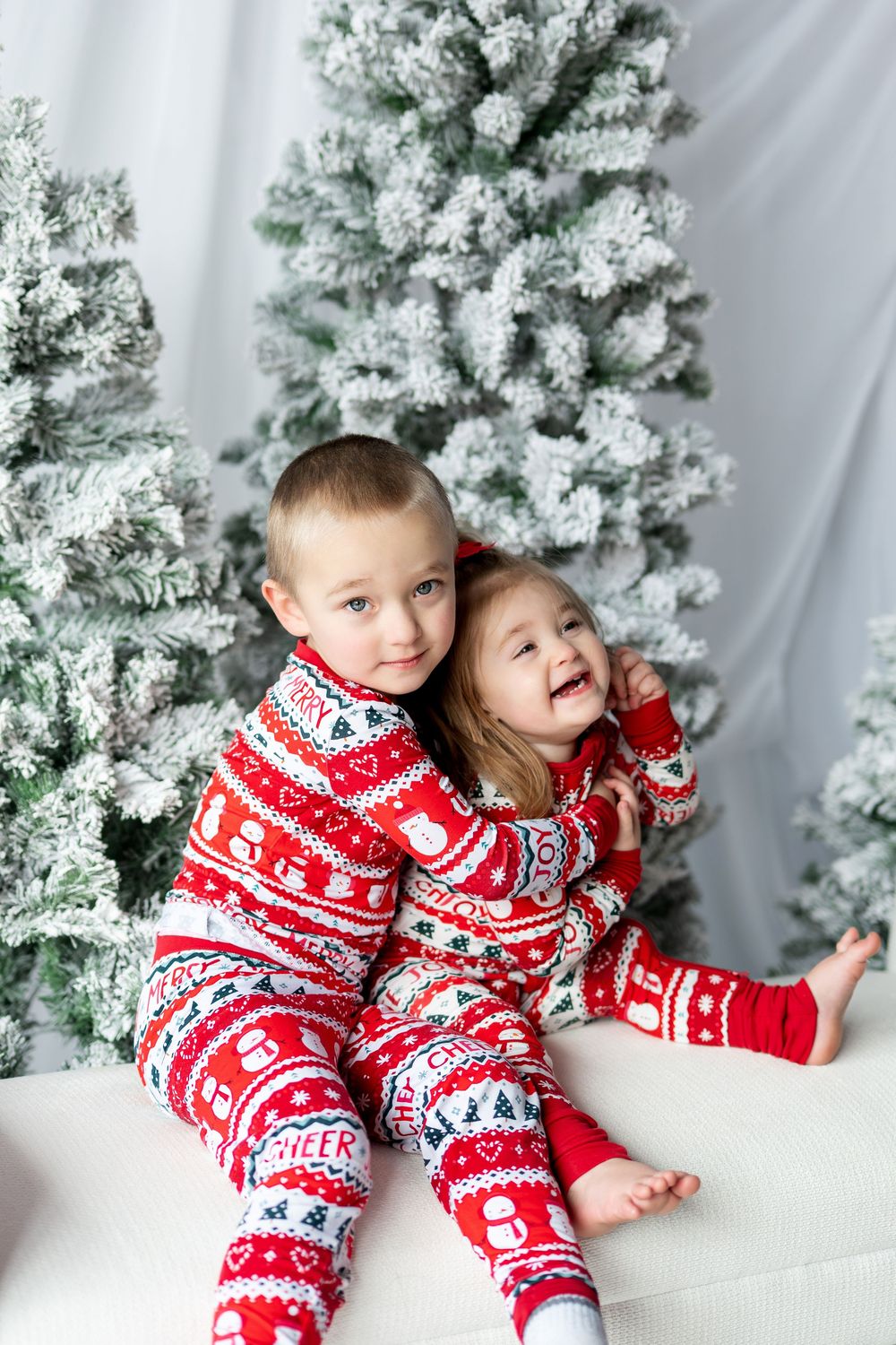 A young boy hugs his giggling little sister, both dressed in matching red Christmas pajamas by snowy trees.