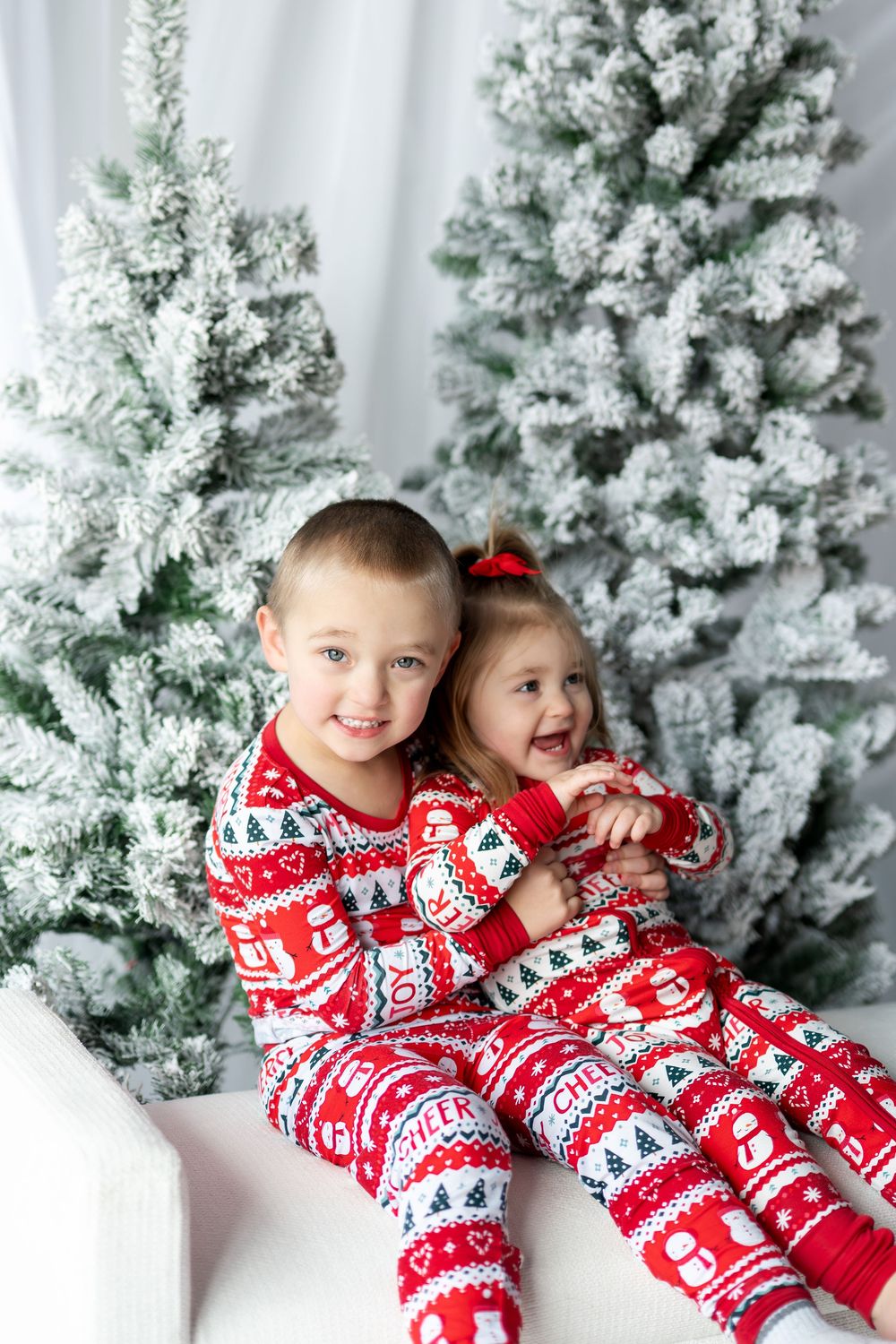 Two young siblings in matching red Christmas pajamas smile together in front of snow-flocked holiday trees.