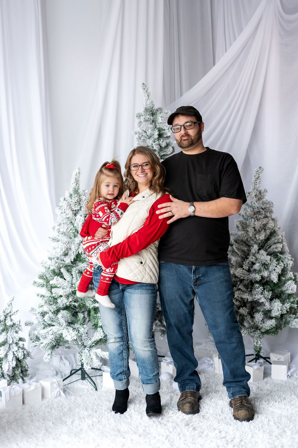 Family of three standing together for a Christmas photo surrounded by snow-flocked trees and gift boxes.