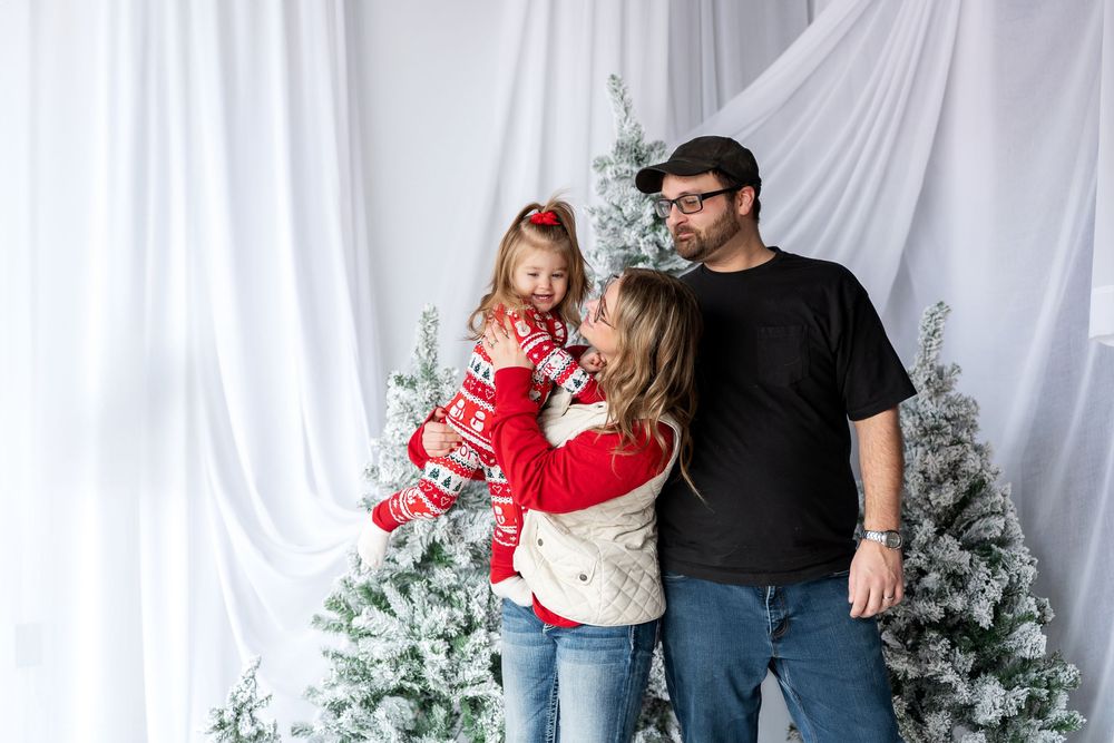 Parents with young daughter in red holiday outfit sharing a joyful moment near snow-flocked Christmas trees.