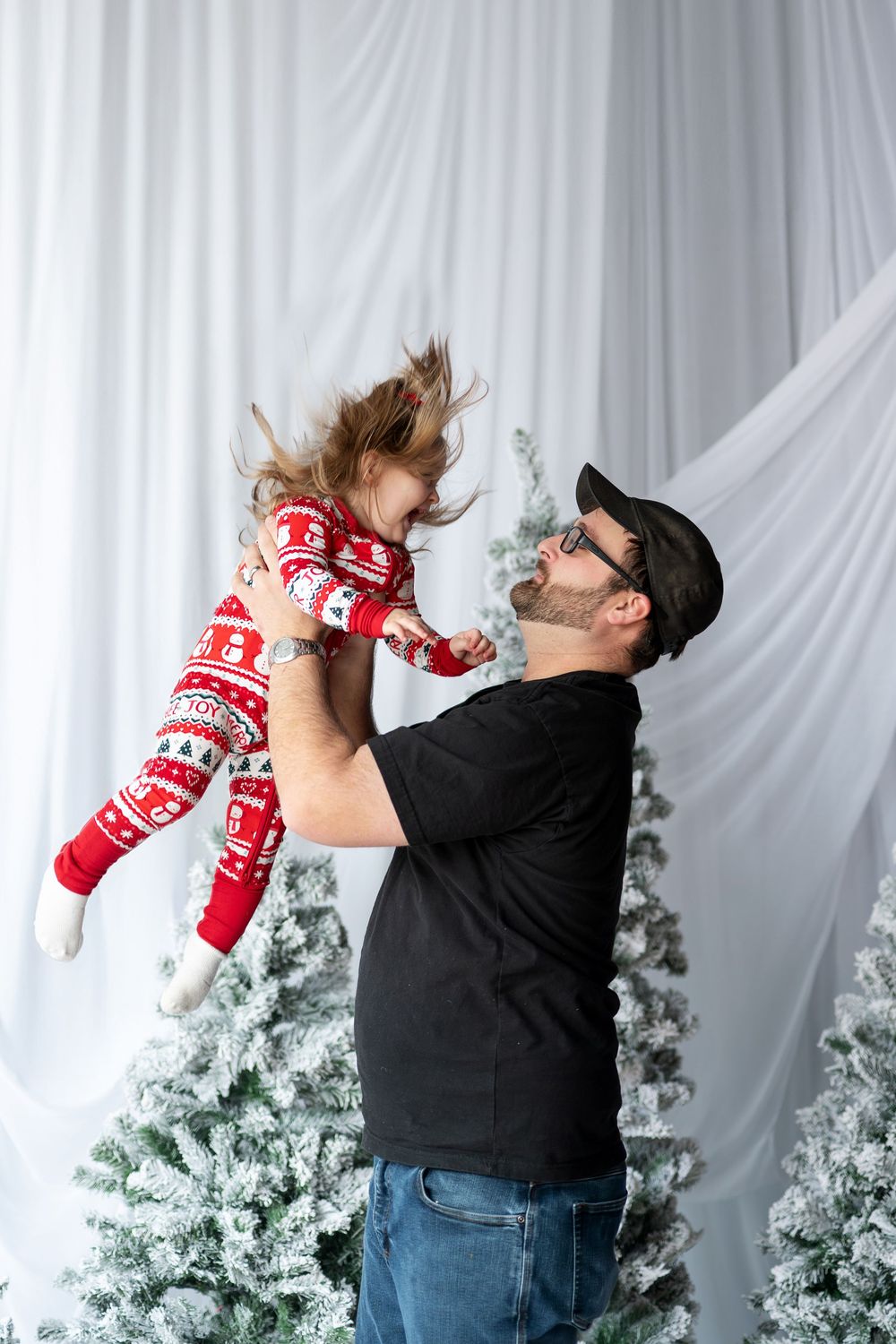 Playful father lifting his laughing daughter in red Christmas pajamas high in the air during holiday photo session.