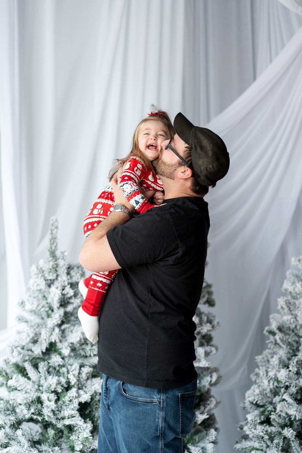 Father kissing laughing toddler girl in red Christmas pajamas near snowy trees.