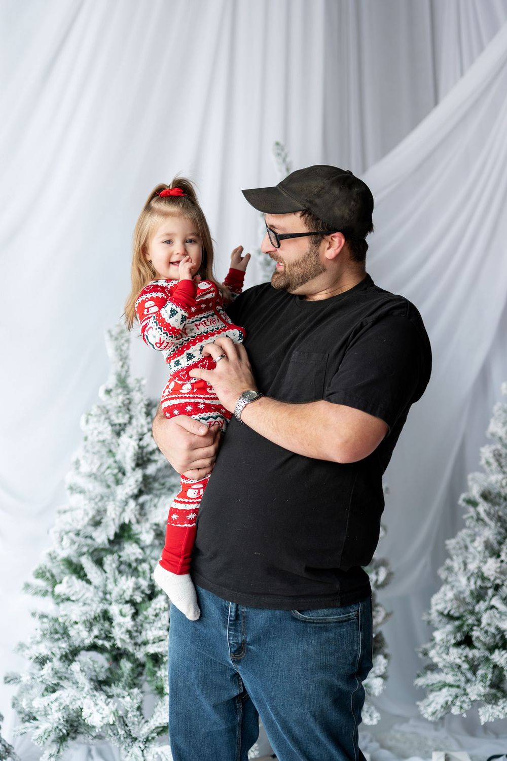 Dad holding smiling little girl in red holiday onesie by flocked Christmas trees.