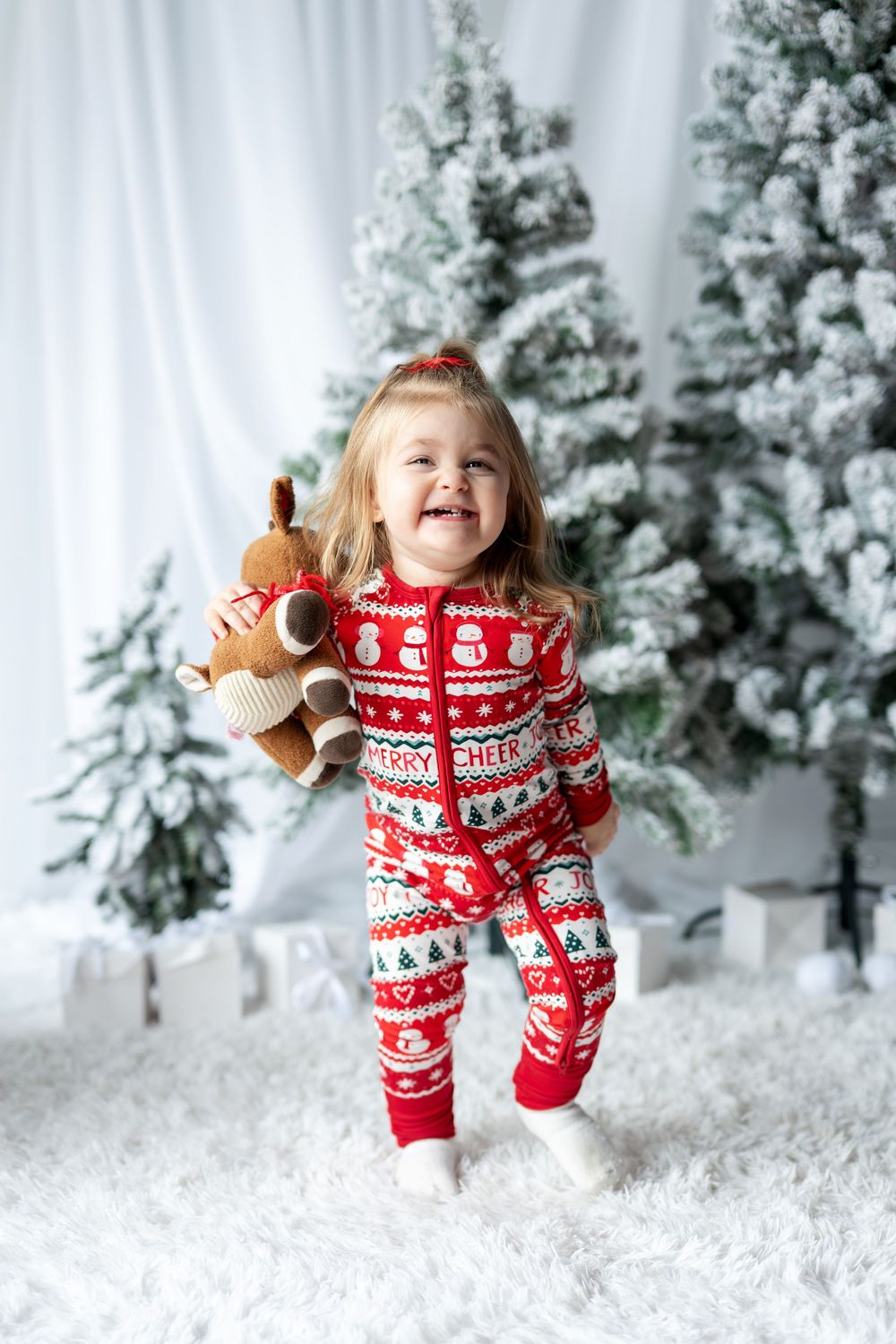 Happy toddler girl in red Christmas onesie holding reindeer stuffed animal by trees.