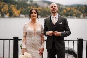 A couple in wedding attire stands by a lakeside railing during an autumn day with fall foliage in the background.