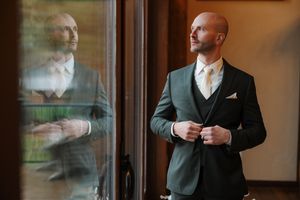 A person in a dark suit adjusts their jacket cuff while standing near a window with reflective glass panels.