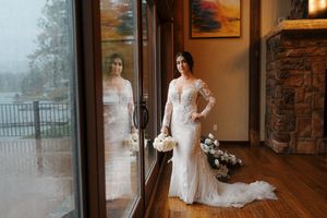 A bride in a white wedding dress stands near a window in an elegant room with warm lighting and stone walls.