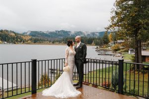 A couple shares an intimate moment on a scenic balcony overlooking a lake and mountains on their wedding day.