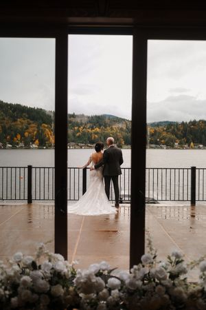 A couple stands on a rainy deck overlooking a misty lake with autumn trees in the background.
