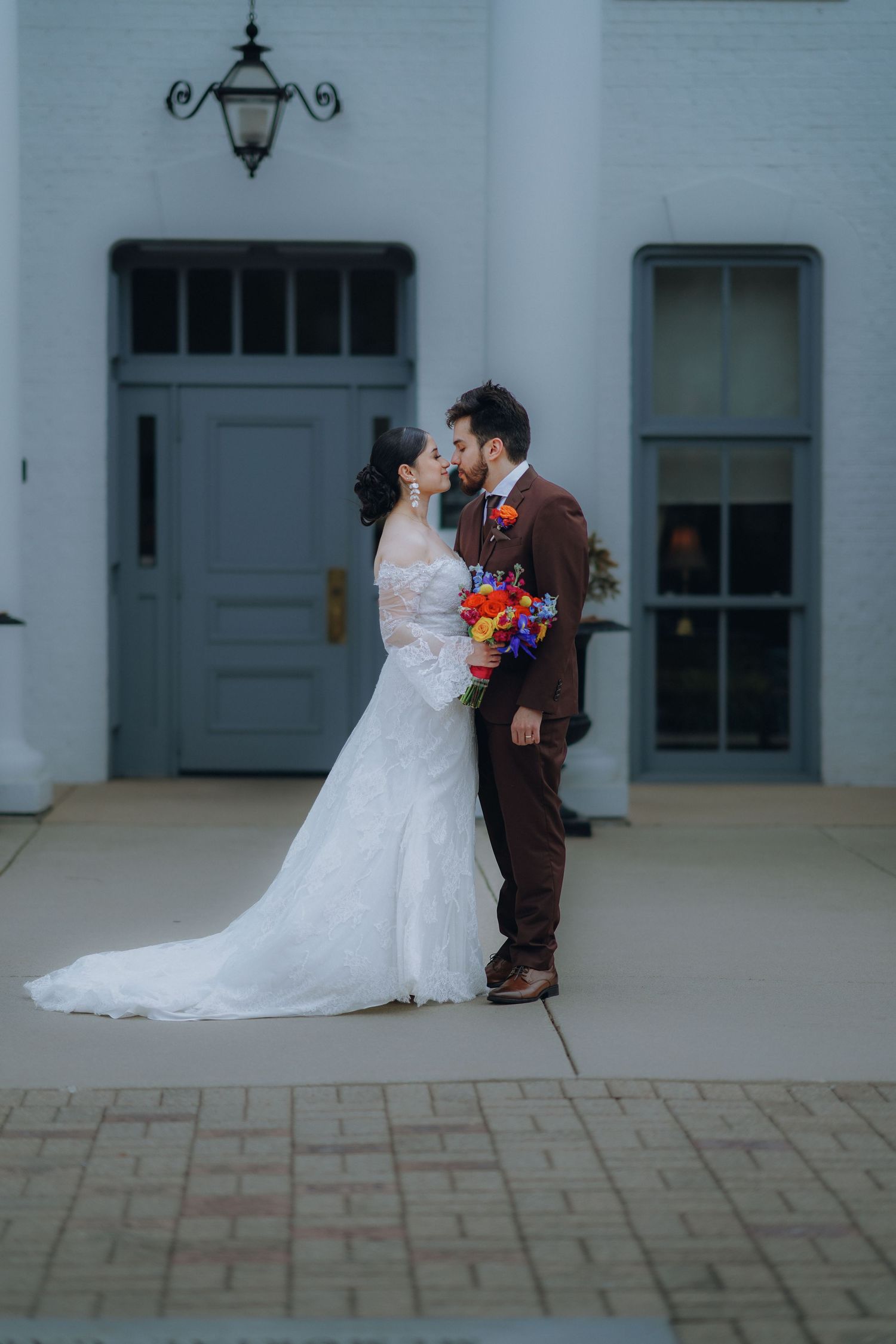 A romantic wedding couple embraces in front of a light blue building with a vintage lantern and door.