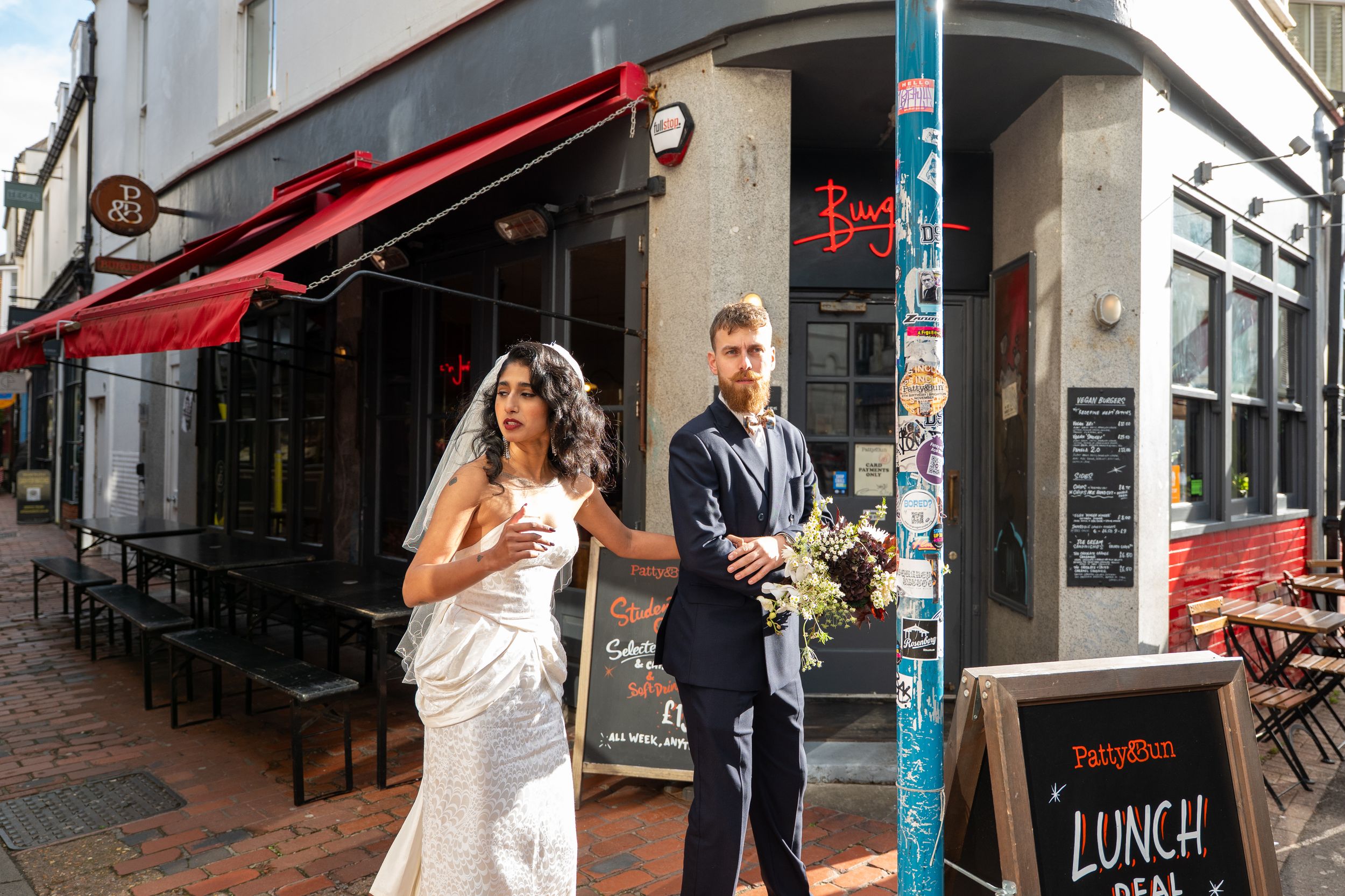 A couple in wedding attire walks past a restaurant with a red awning and outdoor seating on a city street corner.