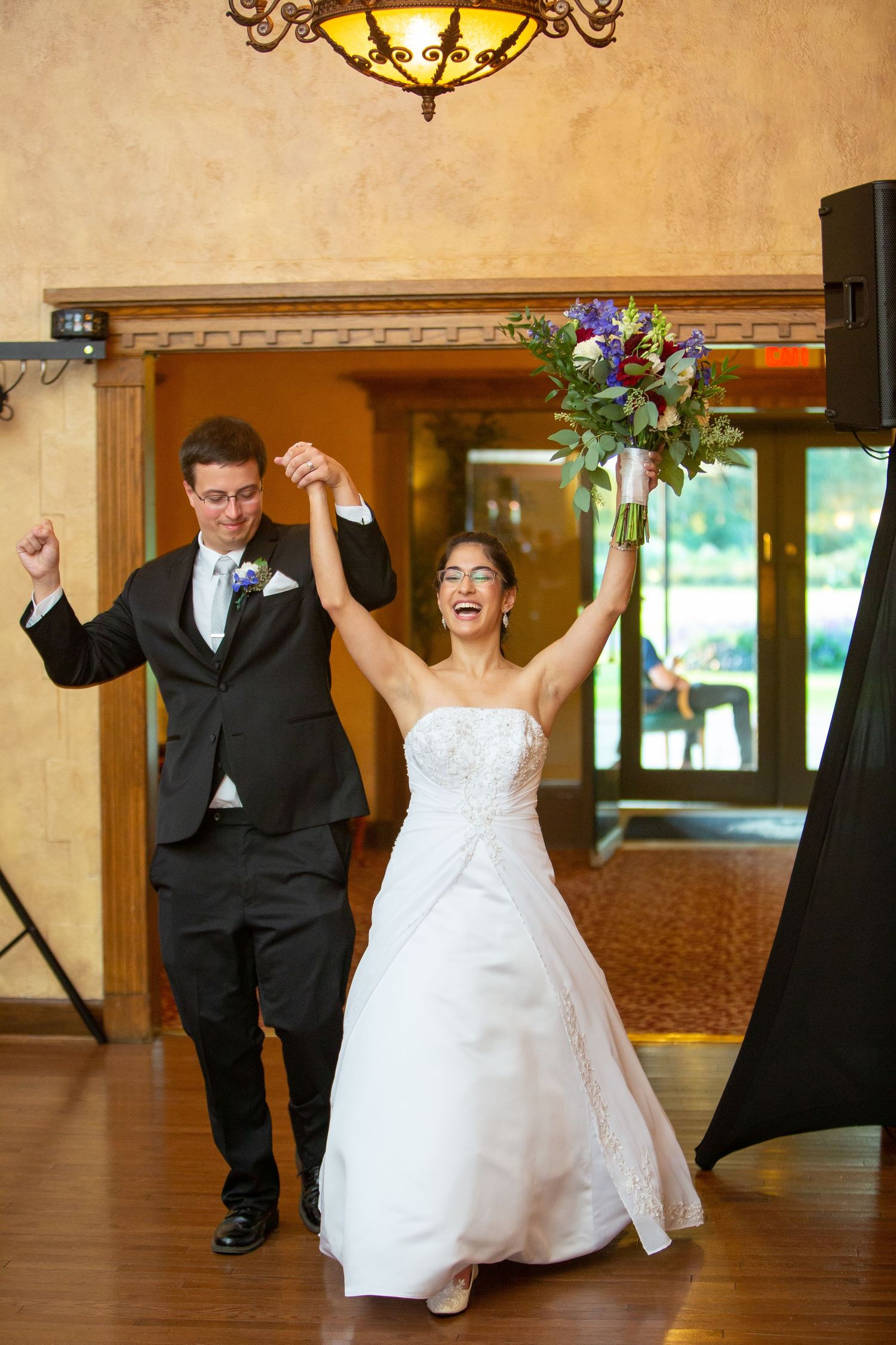 A bride and groom dance joyfully in a grand ballroom with warm lighting and wooden floors.