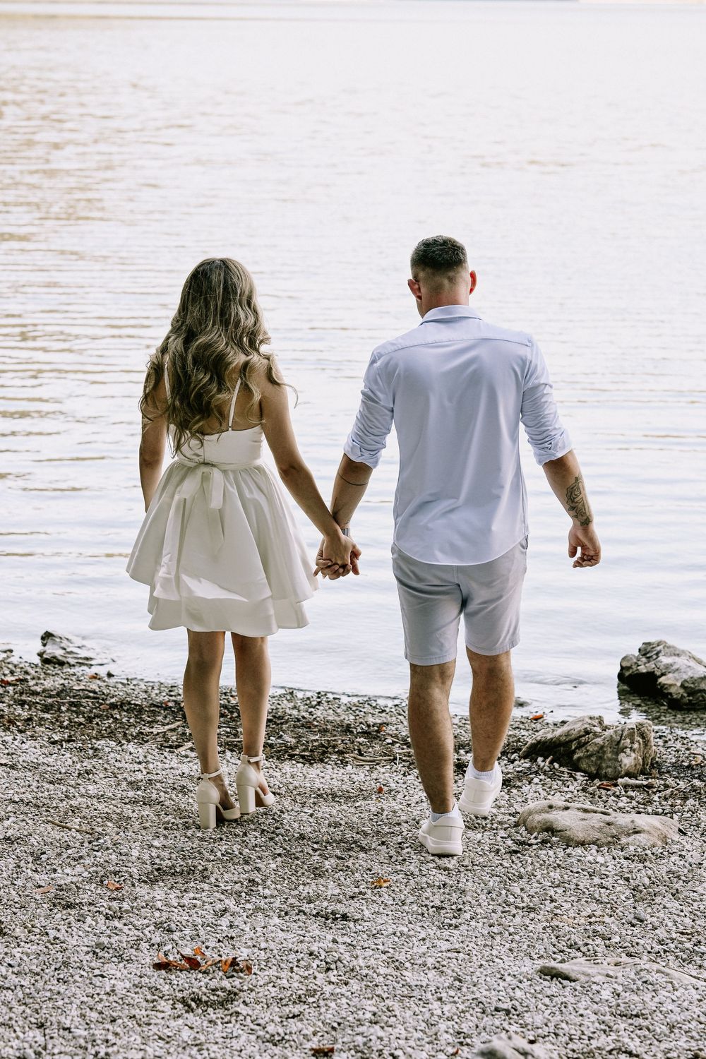 Couple in white outfits walks together on rocky beach shoreline.
