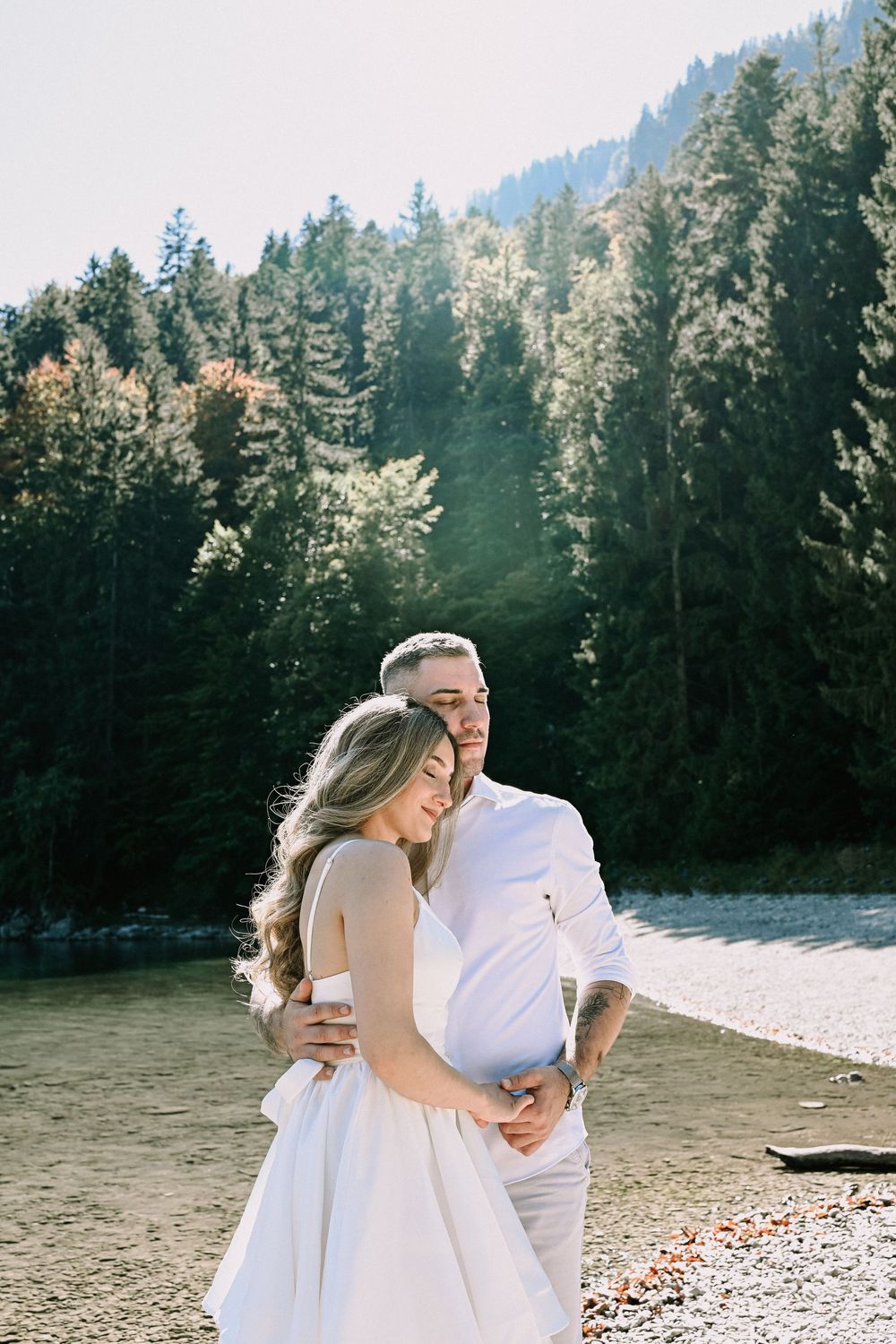 Intimate moment between couple in white clothing on beach with pine forest backdrop.
