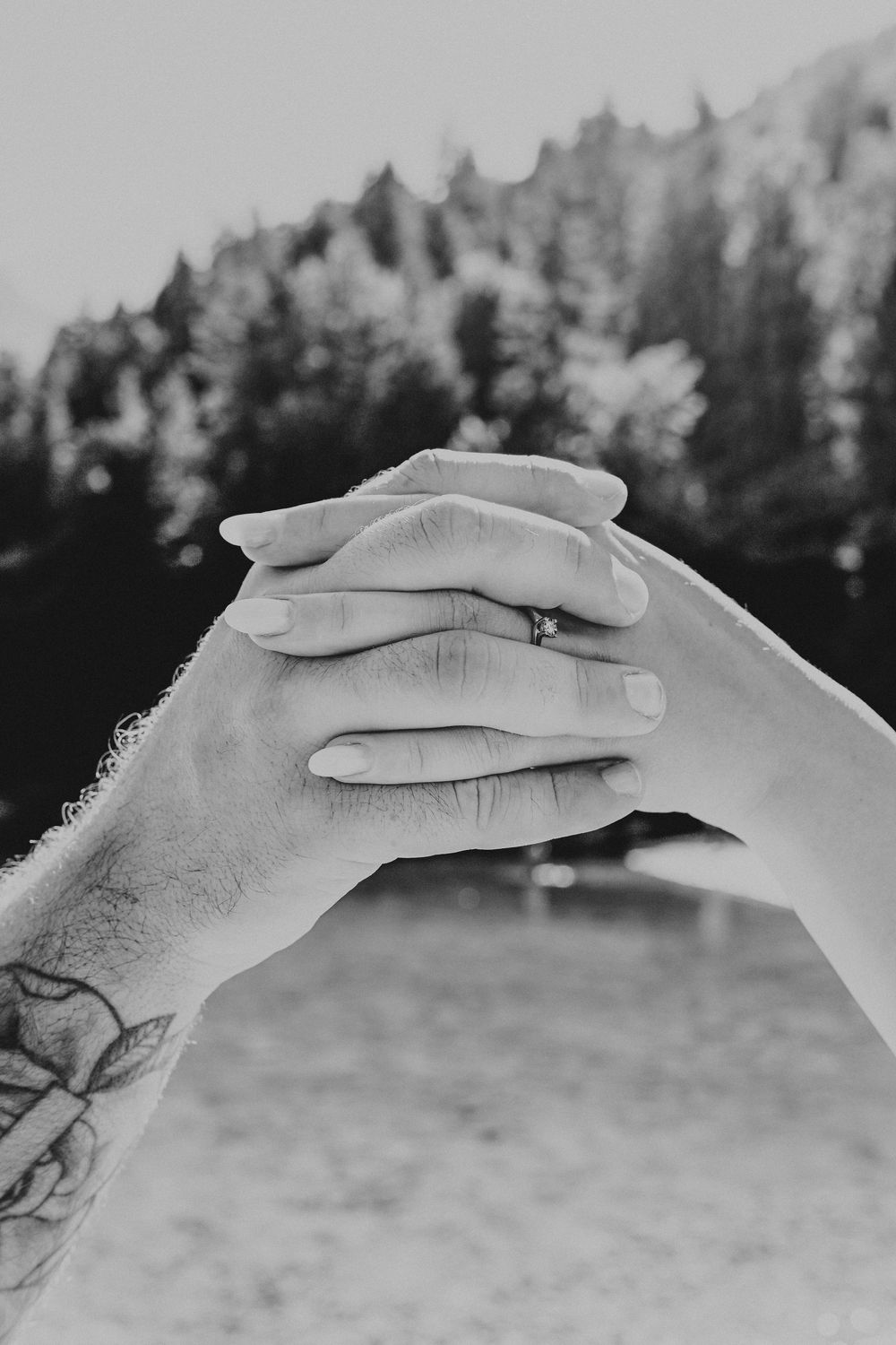 Close-up black and white photo of hands with tattoos clasped together against a forested background.