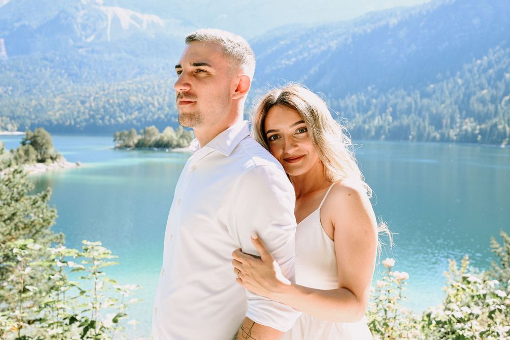 A couple in white outfits poses by a turquoise mountain lake on a sunny day.