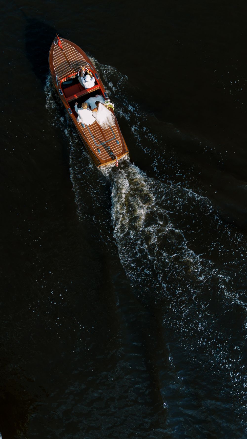 Aerial view of a wooden speedboat cutting through dark water creating dramatic white wake patterns. NC WEDDING PHOTOGRAPHER