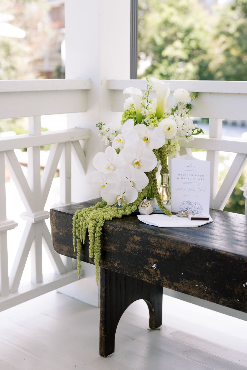White floral arrangements with cascading greenery decorate a rustic dark wood bench on an elegant white porch.NC WEDDING PHOTOGRAPHER