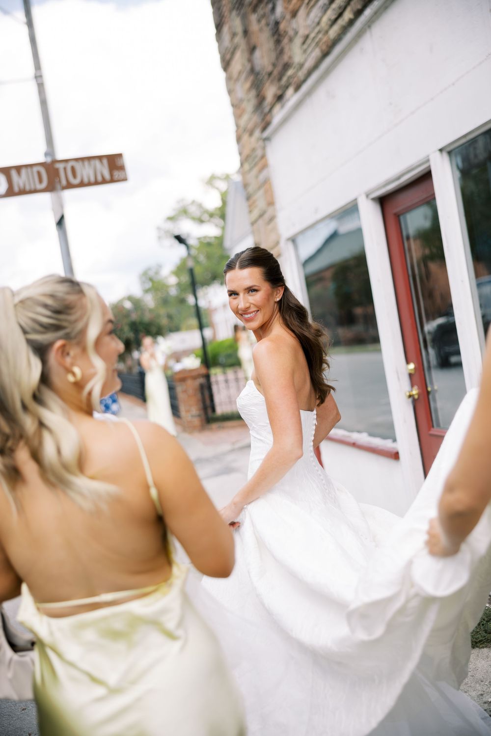 A bride in a white wedding dress smiles over her shoulder while her dress is held by someone in a light colored gown.NC WEDDING PHOTOGRAPHER