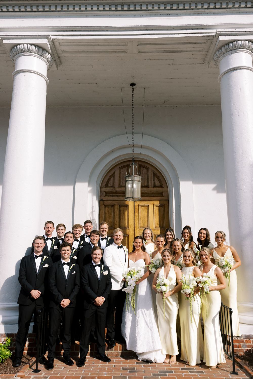 A large wedding party poses together on the steps of a grand white columned building entrance.NC WEDDING PHOTOGRAPHER