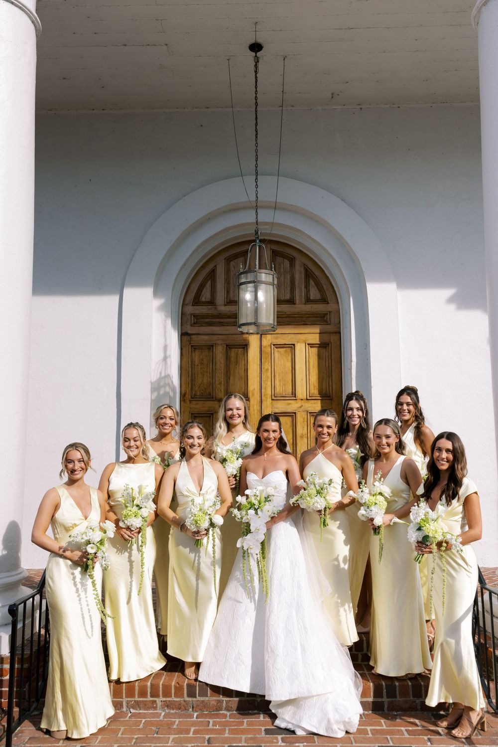 A wedding party in pale yellow dresses poses together on a white columned porch with arched wooden doors.NC WEDDING PHOTOGRAPHER