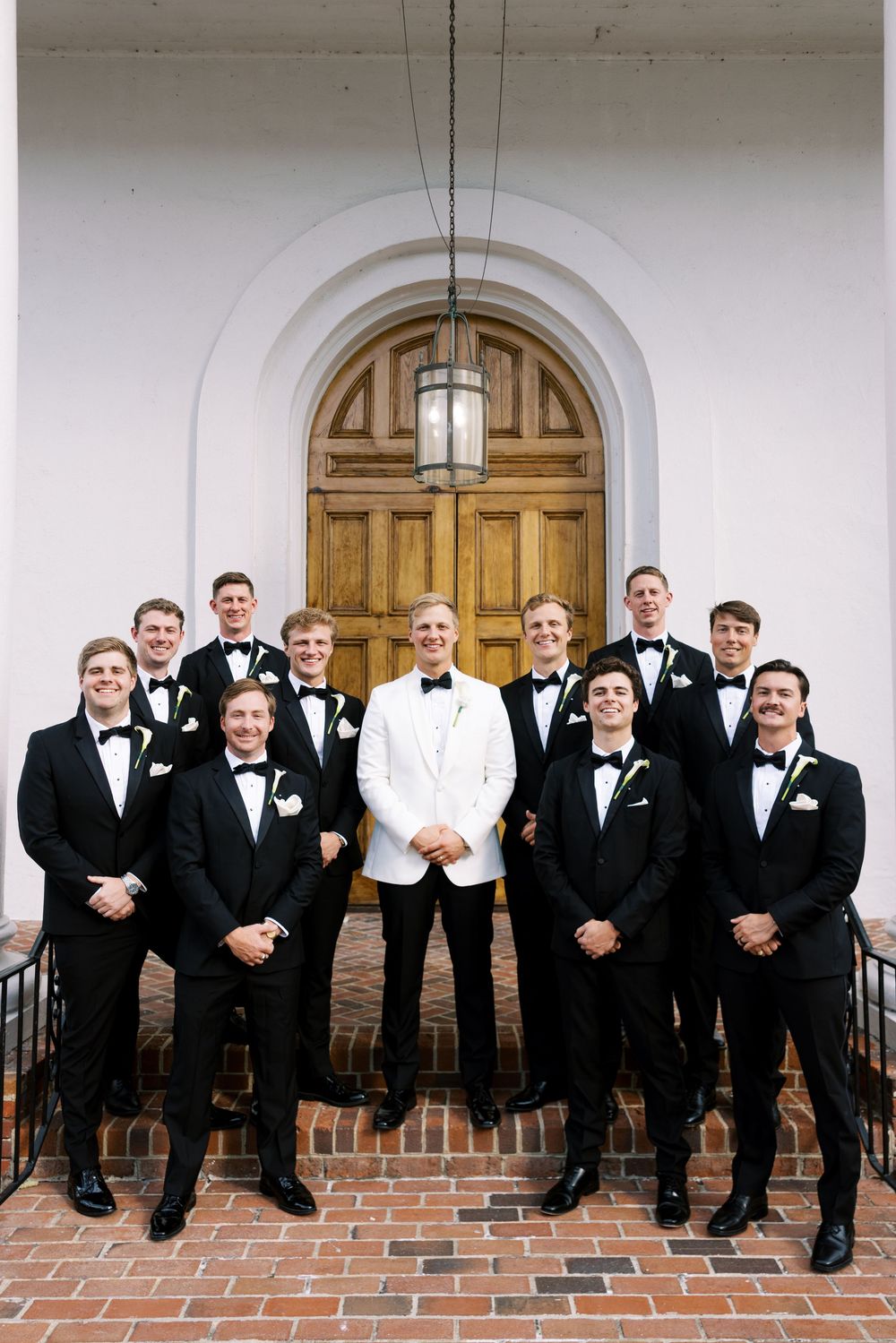 Groomsmen in black tuxedos and bowties sit casually on brick steps in front of white columns and wooden doors.NC WEDDING PHOTOGRAPHER