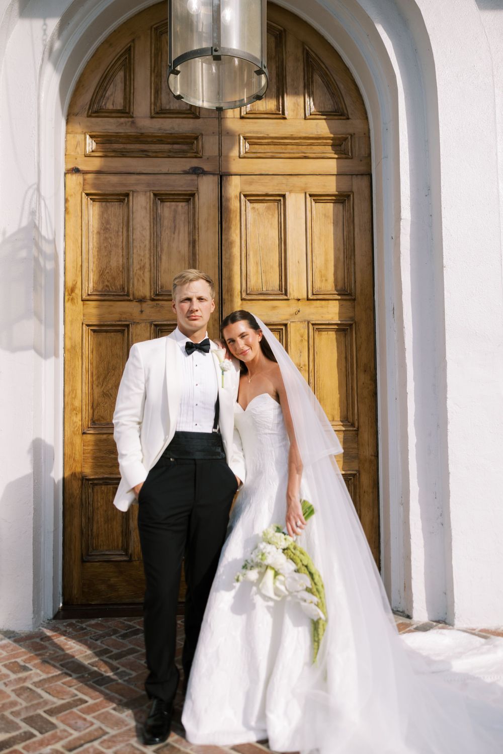 A bride and groom pose in front of ornate wooden doors at their wedding venue with white arched architecture.NC WEDDING PHOTOGRAPHER