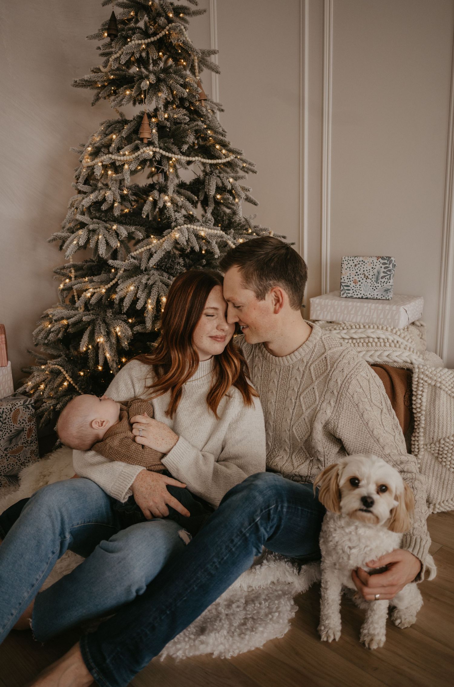 A cozy family scene with a couple, baby, and small white dog sitting by a decorated Christmas tree in warm lighting.