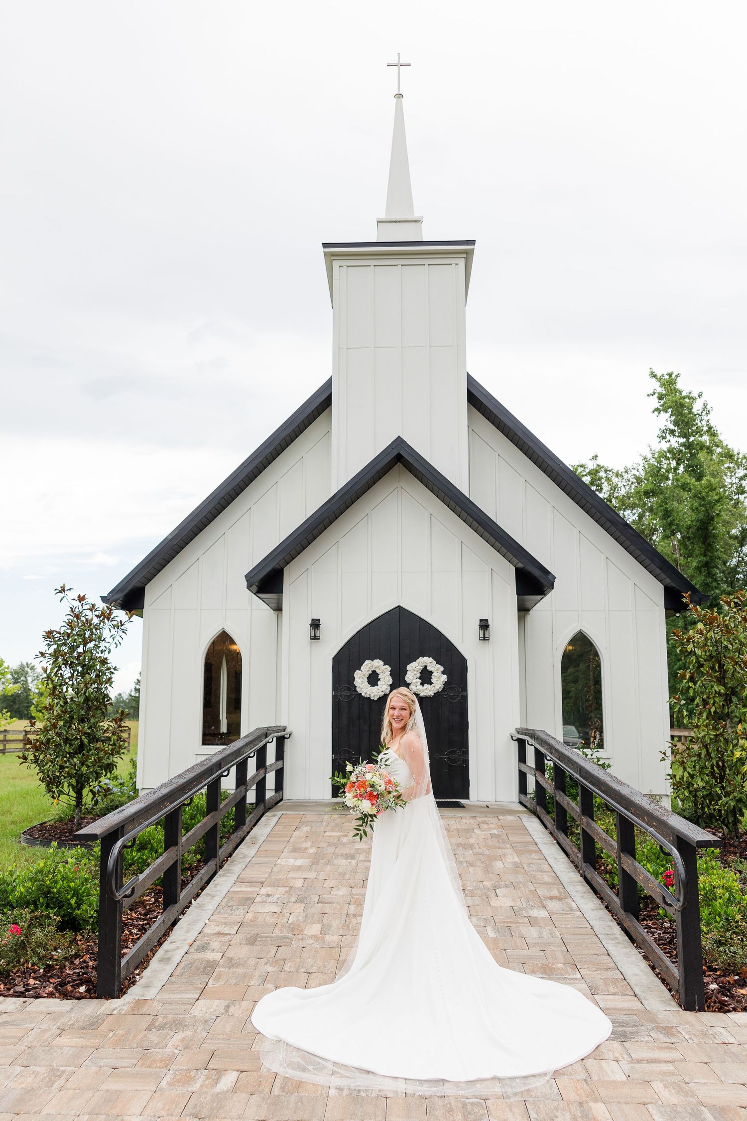 A small white chapel with a steeple sits on manicured grounds surrounded by green trees on a cloudy day.