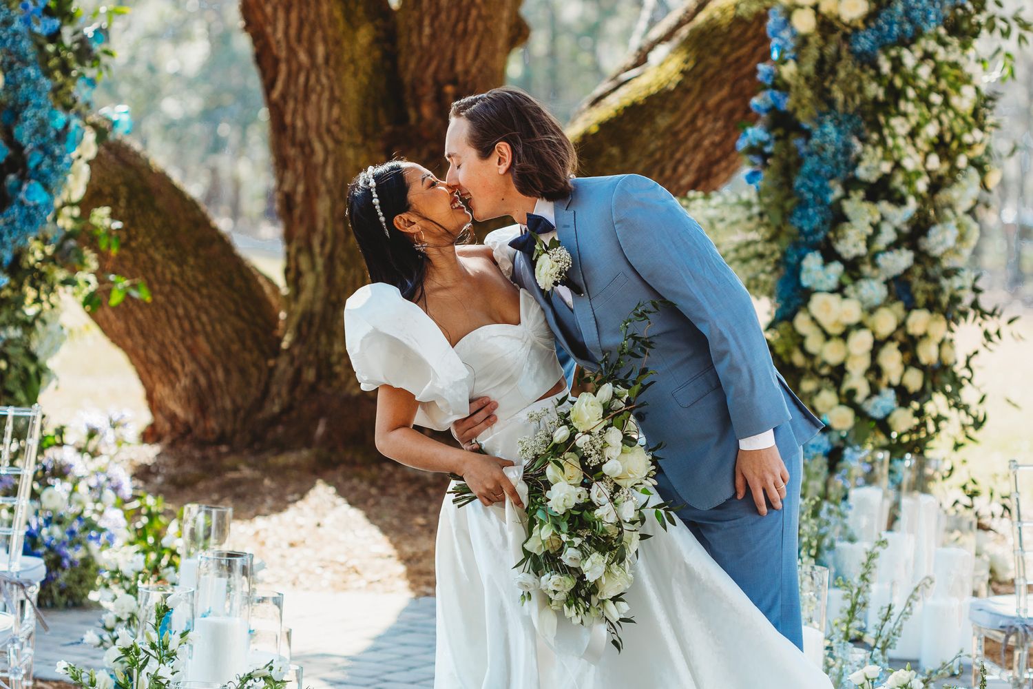 Wedding couple shares romantic moments under a flower-decorated tree arch.