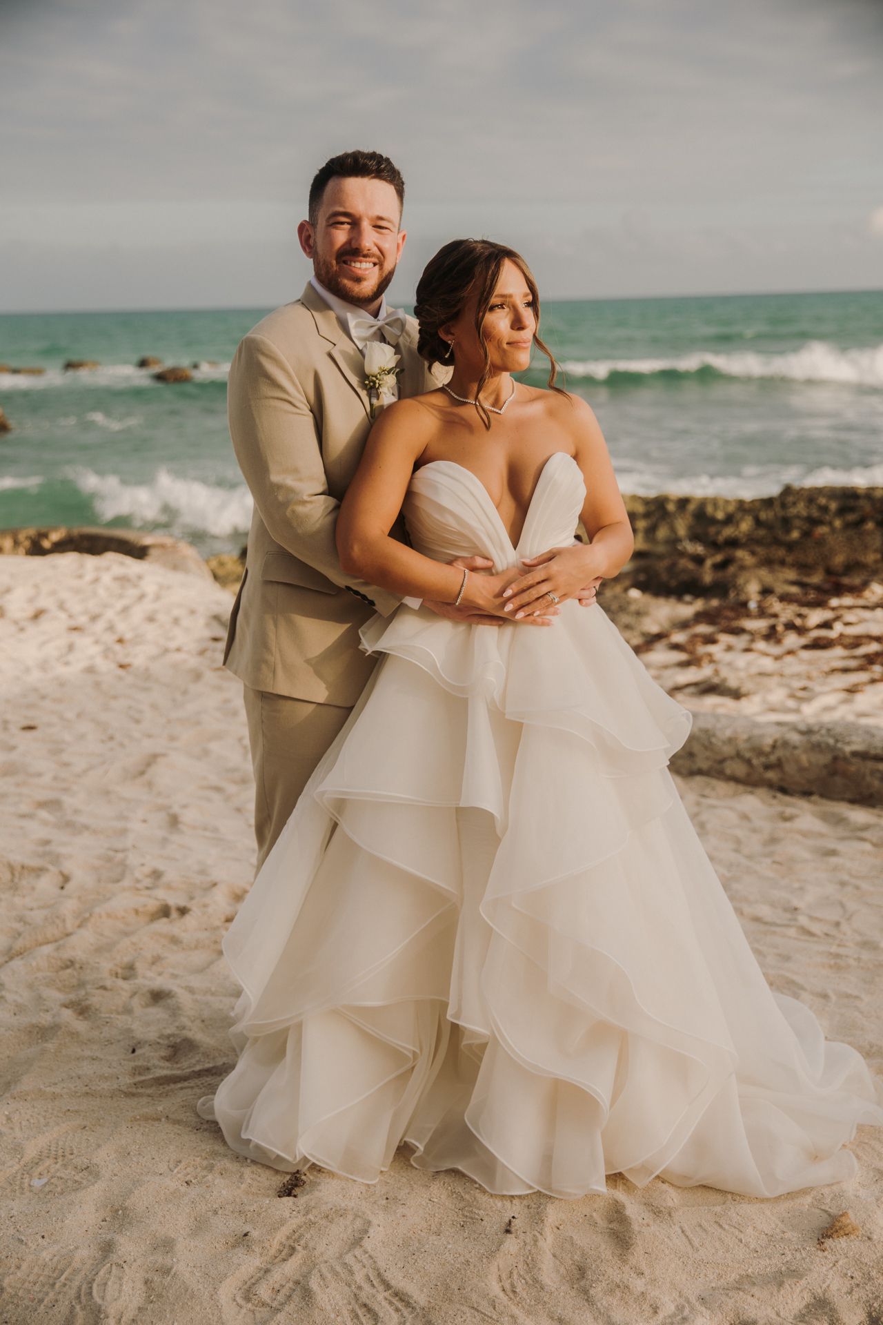 A couple in wedding attire embraces on a scenic beach with waves and rocks in the background. El Dorado Royale Wedding Photographer