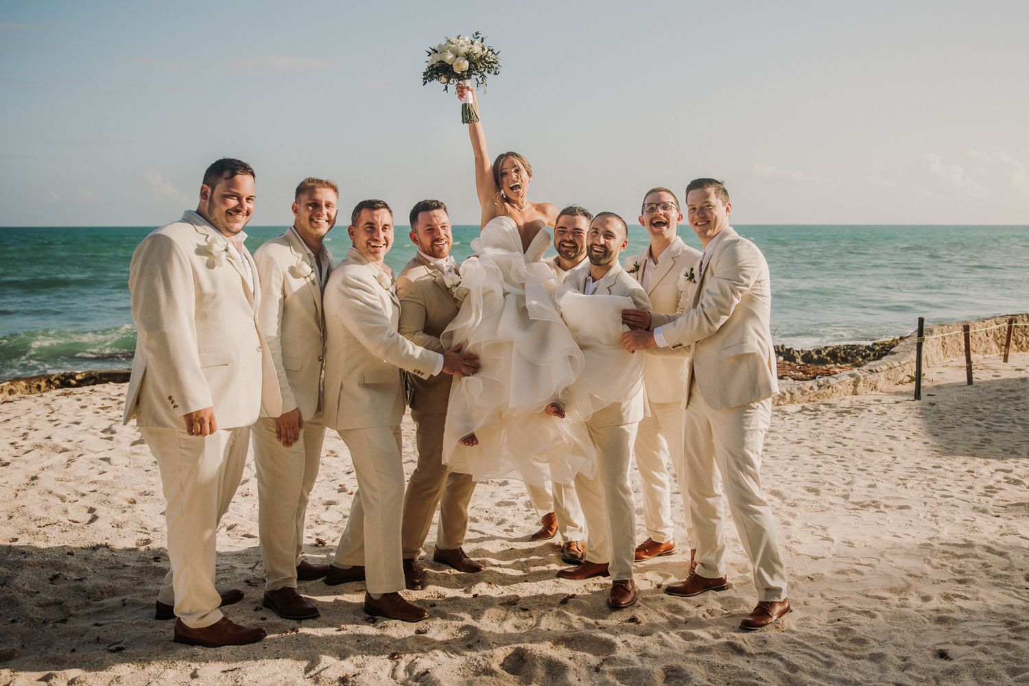 Wedding party in all white attire poses together on a sunny beach with ocean waves in the background. El Dorado Royale Wedding Photographer
